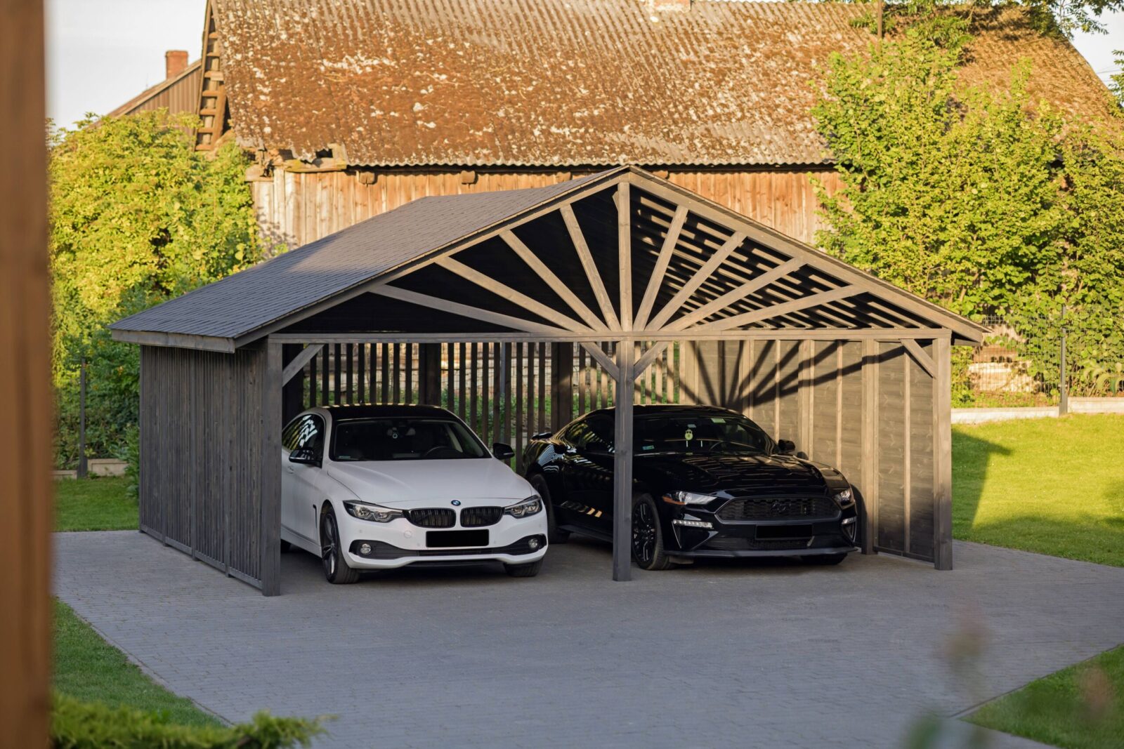 Białe BMW 420 D oraz czarny Ford Mustang GT zaparkowane w nowoczesnej wiacie garażowej w kolorze grafitowym na tle starego drewnianego domu